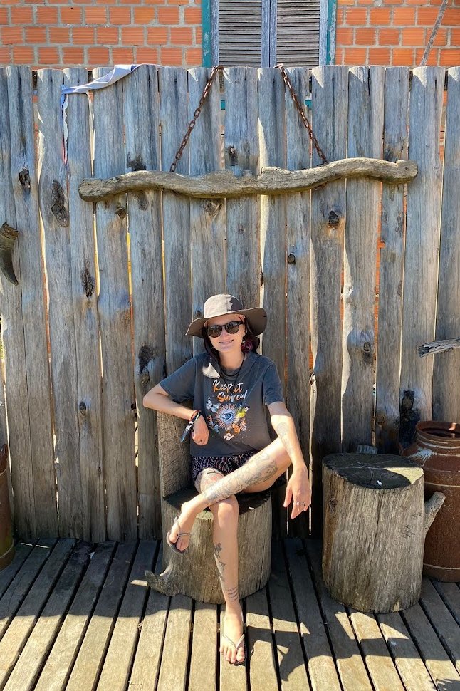 Jaqueline psicóloga de chapéu, sentada em banco rústico, em momento de descanso e conexão com a natureza, representando o autocuidado e o equilíbrio.