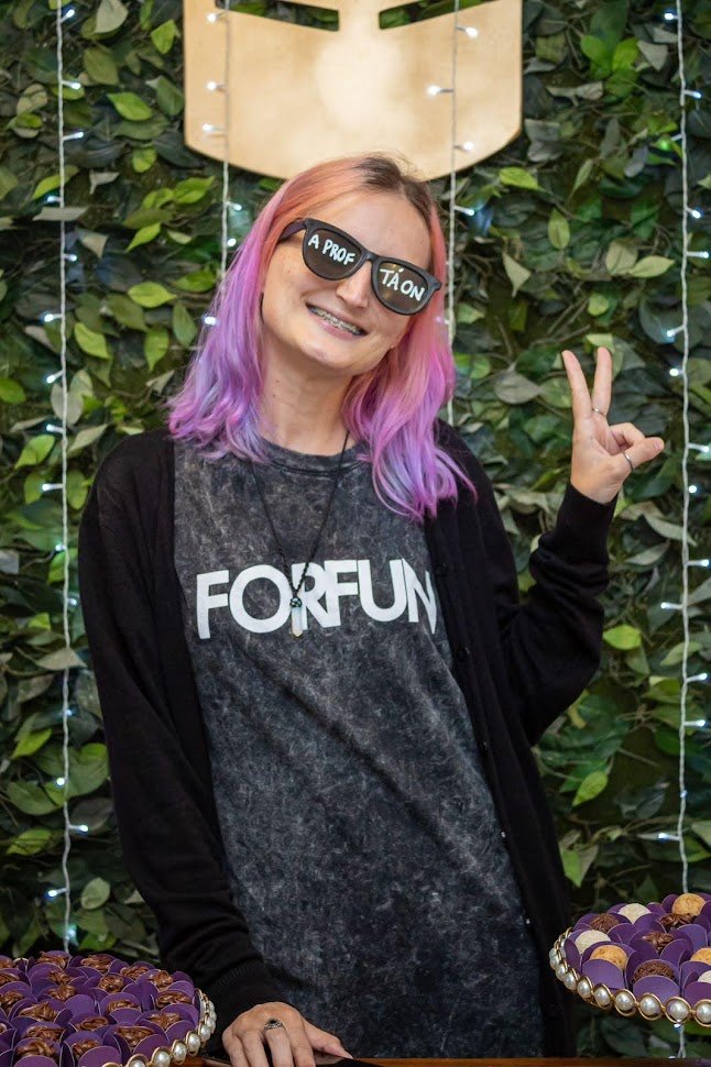 Jaqueline psicóloga sorrindo e fazendo sinal de paz, usando óculos "A Prof. Tá On", transmitindo leveza e bom humor no ambiente profissional.