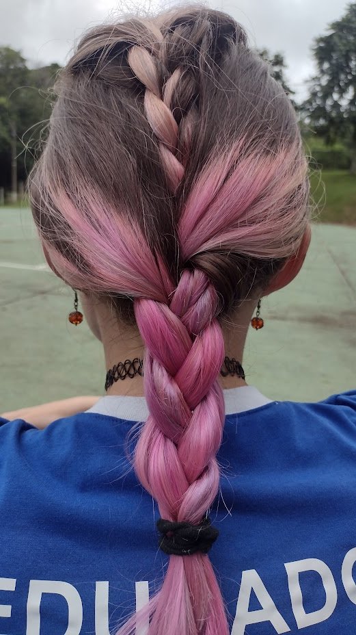 Jaqueline psicóloga de costas com trança rosa, usando camisa de educador, simbolizando a expressão da identidade e autenticidade.